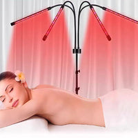 A woman lies face down on a white treatment table under two black LED red-light therapy arms casting pink-red light beams upward, with a white towel wrapped around her body.