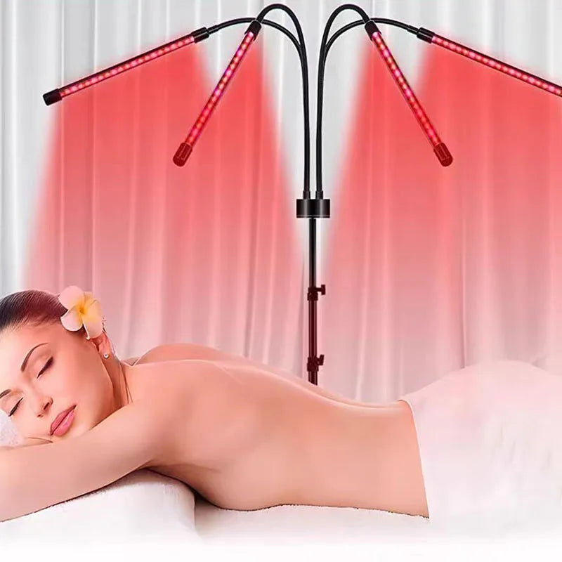 A woman lies face down on a white treatment table under two black LED red-light therapy arms casting pink-red light beams upward, with a white towel wrapped around her body.